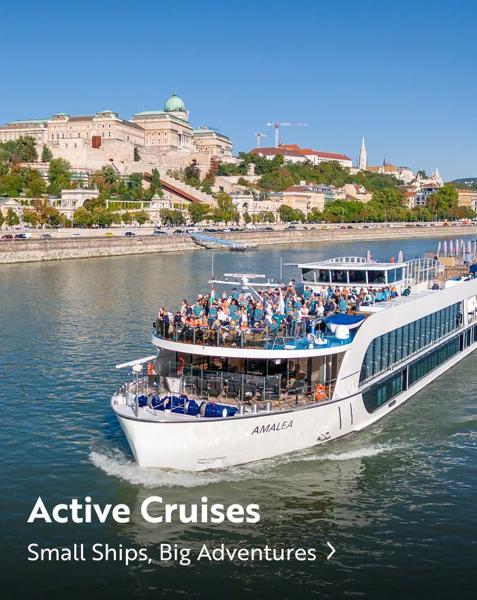 An Amalea river cruise ship passing through Budapest, Hungary, with the historic Buda Castle visible in the background. Text overlay reads: "Active Cruises. Small Ships, Big Adventures".