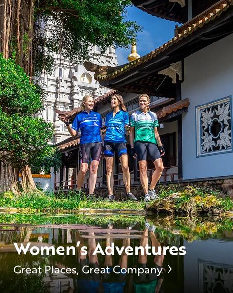 Three women in cycling jerseys and padded shorts stand together beside a tranquil pond near traditional Eastern-style architecture. A towering, ornate temple rises in the background behind lush green foliage. Their reflections are mirrored in the clear water below. White text overlaid near the bottom reads "Women's Adventures" followed by "Great Places, Great Company >".