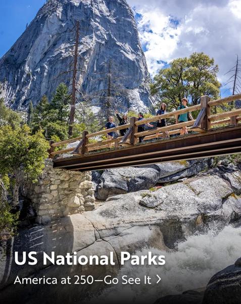 A group of people standing on a wooden bridge over a waterfall with a large rock mountain in the background at a US National Park. Text overlay reads: "US National Parks. America at 250&mdash;Go See It".