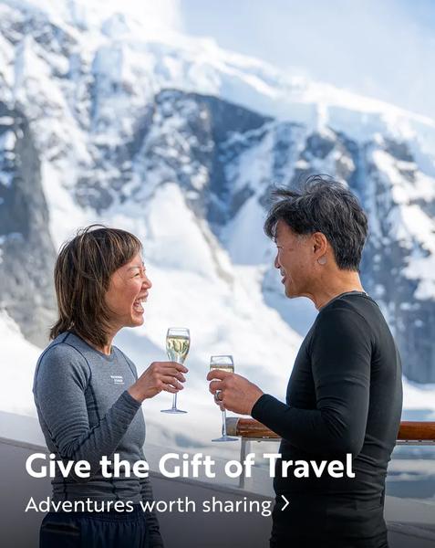A smiling couple stands on the deck of a ship, holding glasses of champagne and facing each other against a backdrop of a massive, snow-covered mountain. Text at the bottom reads, "Give the Gift of Travel. Adventures worth sharing."