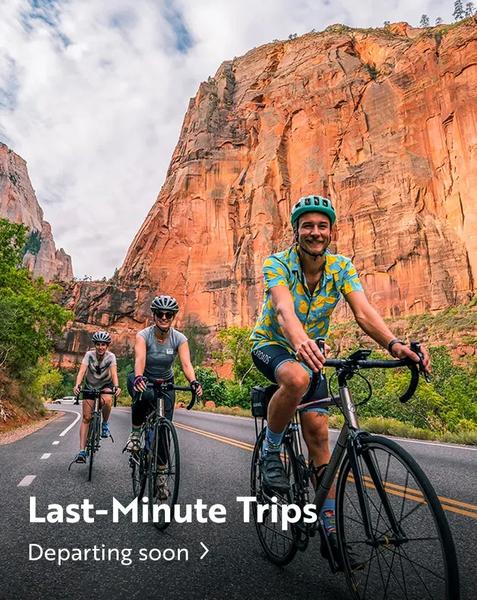 Three cyclists riding road bikes down an asphalt road lined with trees, against a backdrop of massive, towering red rock cliffs under a partly cloudy sky. The lead cyclist, a smiling man wearing a blue and yellow patterned shirt and a teal helmet, looks toward the camera. Text overlay at the bottom reads "Last-Minute Trips" and "Departing soon >".