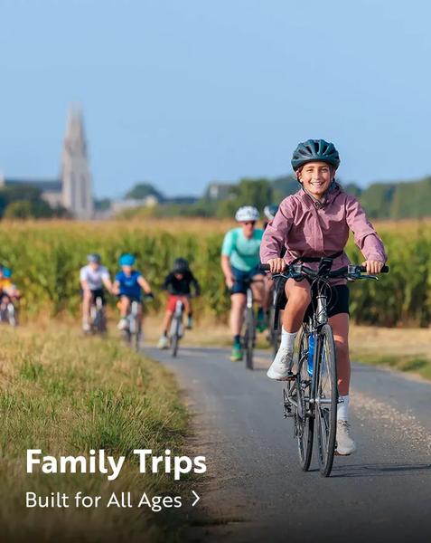 A young girl smiling and riding a bicycle down a paved path along a field, leading a group of cyclists including children and adults wearing helmets. In the distance, a tall church spire rises above the countryside under a clear blue sky. White text overlaid near the bottom left reads "Family Trips" and "Built for All Ages >".