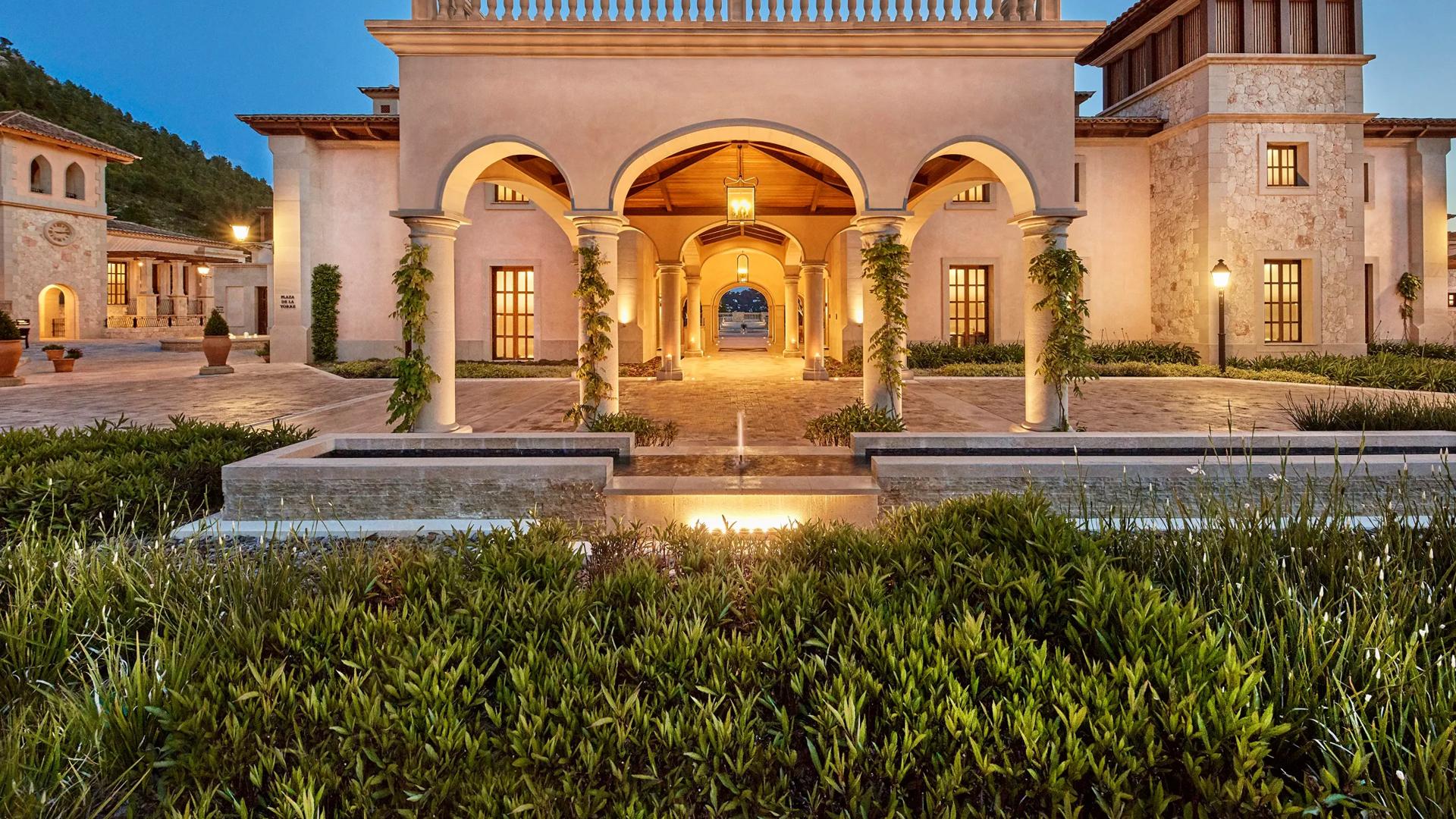 Evening view of an illuminated Cap Vermell Grand Hotel entrance featuring stone arches, columns wrapped in climbing vines, and a paved courtyard under a twilight sky.
