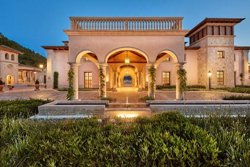 Evening view of an illuminated Cap Vermell Grand Hotel entrance featuring stone arches, columns wrapped in climbing vines, and a paved courtyard under a twilight sky.