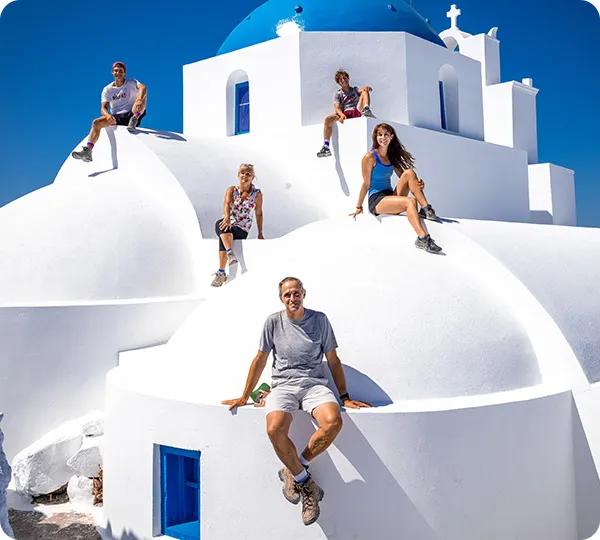 A group of five people posing together while sitting on the white domed roof structure of a traditional blue-domed Greek church under a clear blue sky.
