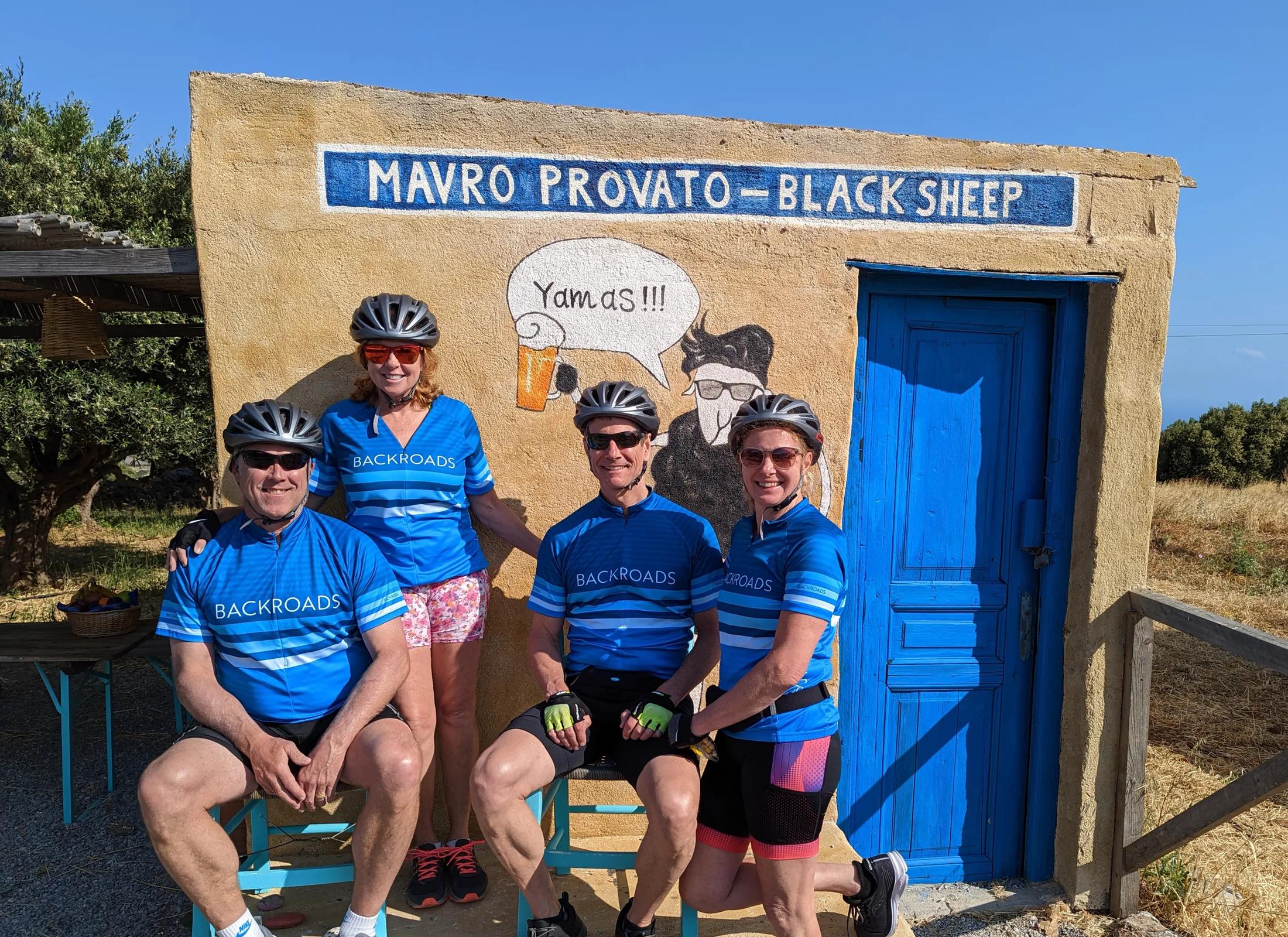 A group of four guests wearing branded Backroads cycling gear, posing in front of a rustic building with a blue door and a mural with sheep drinking beer, labeled "Mavro Provato - Black Sheep" in Crete, Greece.