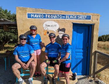 A group of four guests wearing branded Backroads cycling gear, posing in front of a rustic building with a blue door and a mural with sheep drinking beer, labeled "Mavro Provato - Black Sheep" in Crete, Greece.
