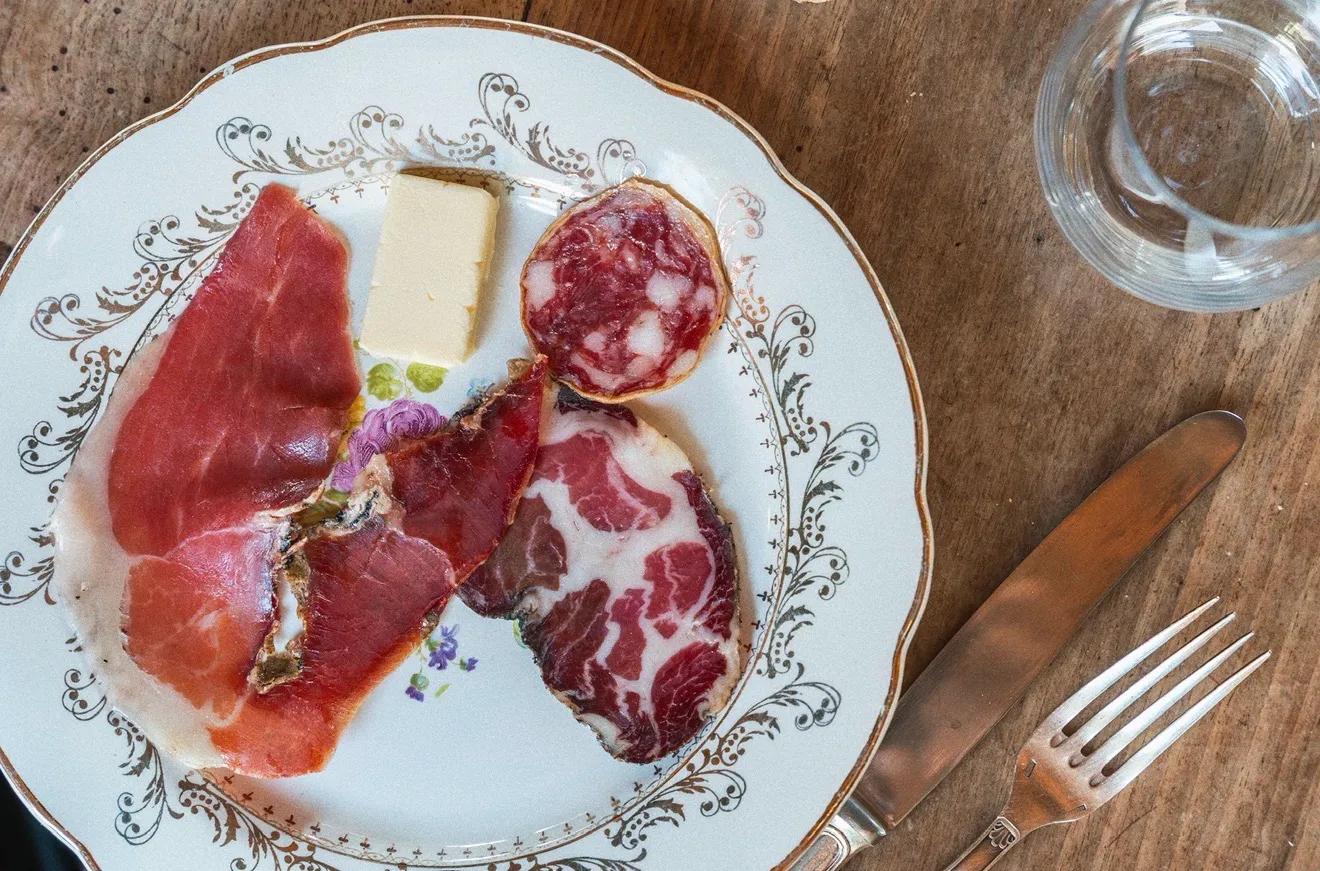 An assortment of cured meats (salumi and ham) and a piece of butter served on an ornate vintage plate with a fork and knife at A Pignata, in Levie, Corsica, France.