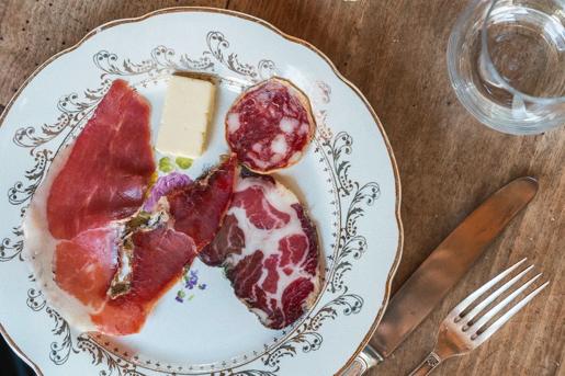 An assortment of cured meats (salumi and ham) and a piece of butter served on an ornate vintage plate with a fork and knife at A Pignata, in Levie, Corsica, France.