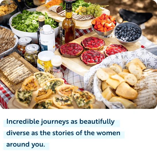 A high-angle view of a colorful outdoor picnic spread over a red-and-white checkered tablecloth. The feast includes bowls of fresh salad, sliced strawberries, and blueberries, wooden boards with red berry tarts, sliced bread, quiche slices, green grapes, cured meats, and various jars and oil bottles. Dark blue text on a white background below the image reads, "Incredible journeys as beautifully diverse as the stories of the women around you."