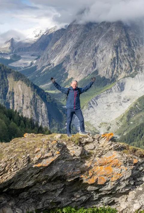 A smiling hiker standing on a rocky ledge with arms raised in celebration, set against a backdrop of dramatic, cloud-capped mountains and alpine valleys.