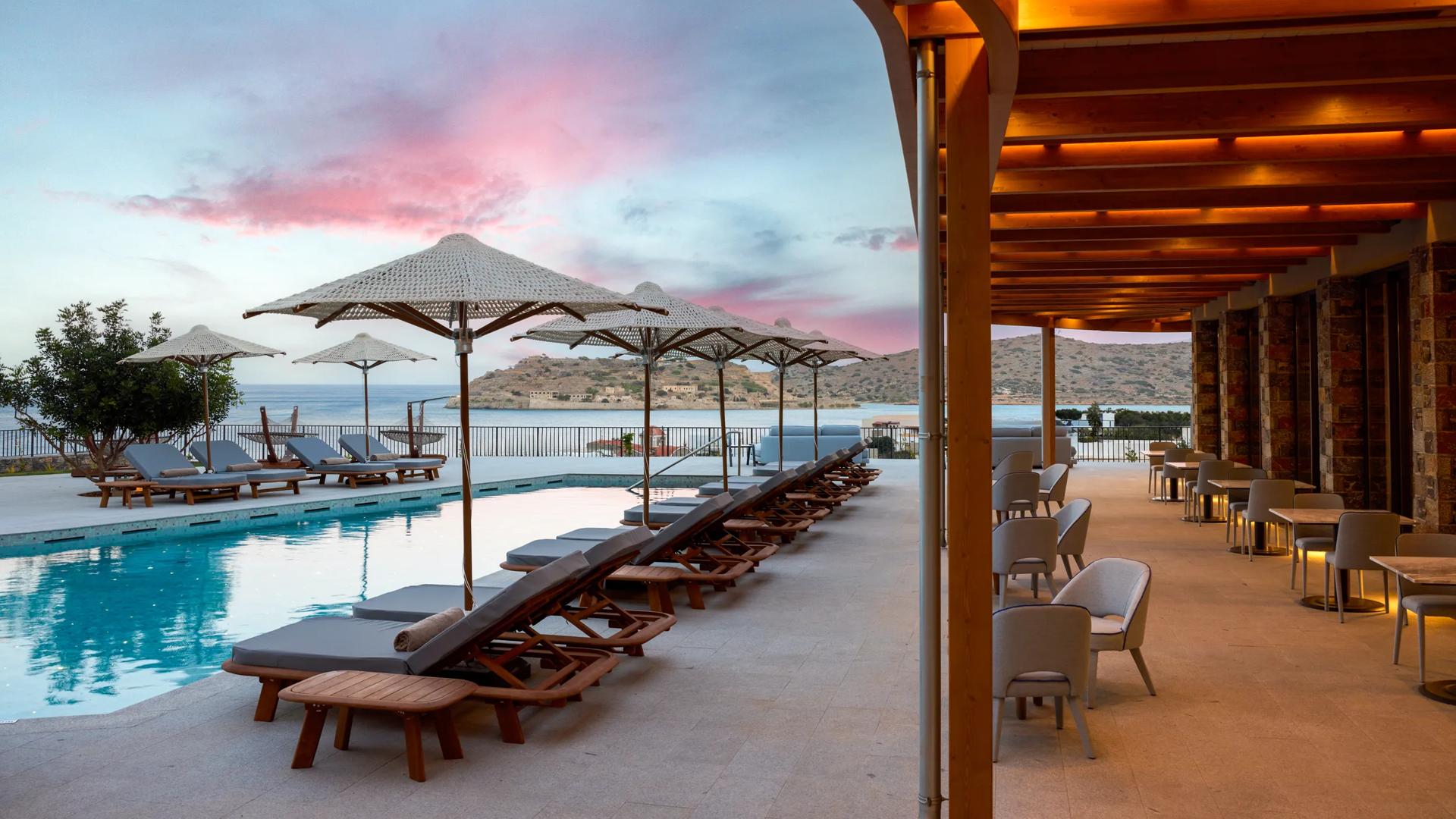 The outdoor pool deck at Cayo Exclusive Resort & Spa in Crete, featuring lounge chairs, umbrellas and covered seating area with a view overlooking Spinalonga Island.