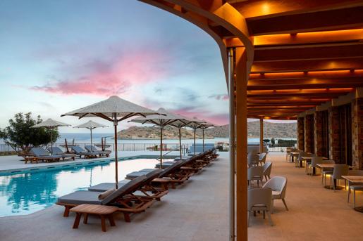 The outdoor pool deck at Cayo Exclusive Resort & Spa in Crete, featuring lounge chairs, umbrellas and covered seating area with a view overlooking Spinalonga Island.