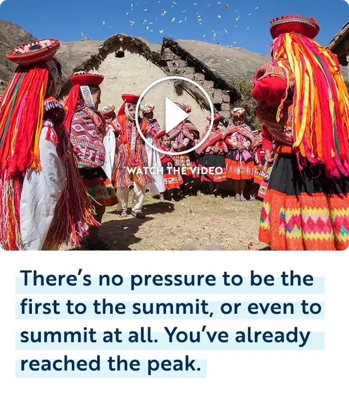A group of people in colorful, traditional Andean clothing dancing in an outdoor courtyard in front of a stone building with mountain peaks in the background, with confetti falling from the sky. A circular video play icon with the text "WATCH THE VIDEO" is overlaid in the center of the image. Below the photo, dark blue text with light blue highlighting reads: "There&rsquo;s no pressure to be the first to the summit, or even to summit at all. You&rsquo;ve already reached the peak."