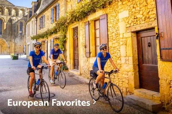 Three cyclists wearing blue shirts and helmets riding bicycles along a cobbled street past historic yellow stone buildings, with text reading "European Favorites" overlaid in the bottom left corner.