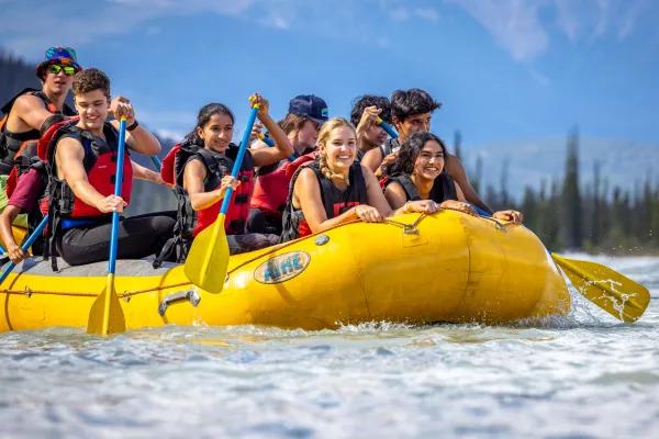 A group of young adults wearing life jackets smiling and paddling together in a yellow inflatable raft on a river, with forested mountains visible in the background.