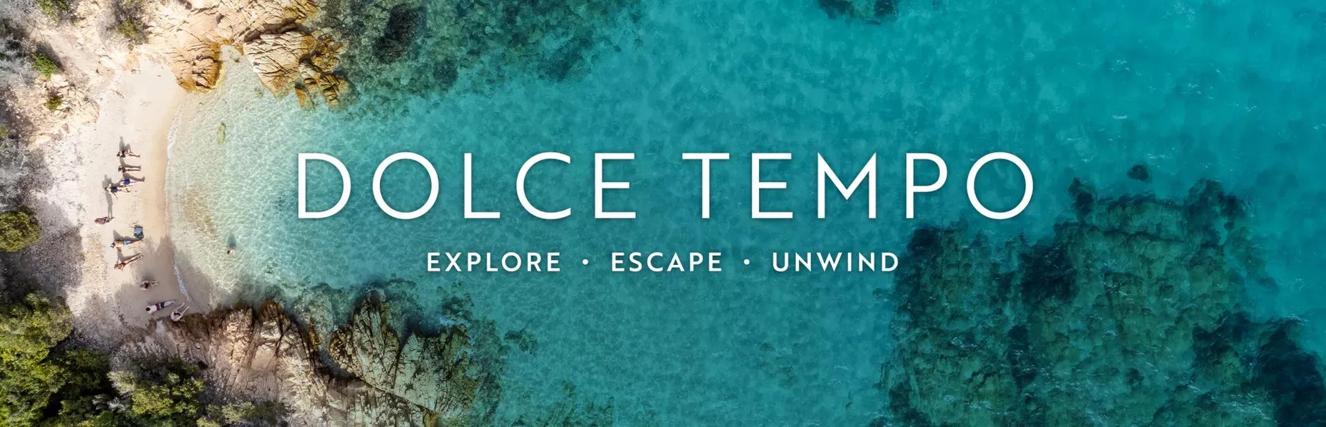 An aerial view of a secluded beach cove with crystal-clear turquoise water, sandy shore, and rocky edges. Overlaid across the center of the image in clean white lettering is the title "DOLCE TEMPO" above the subtitle "EXPLORE • ESCAPE • UNWIND".