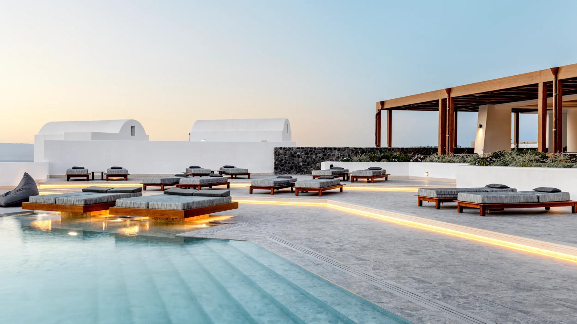 The outdoor pool terrace at the Santo Pure Oia Suites & Villas in Santorini, featuring minimalist wooden loungers and ambient lighting.