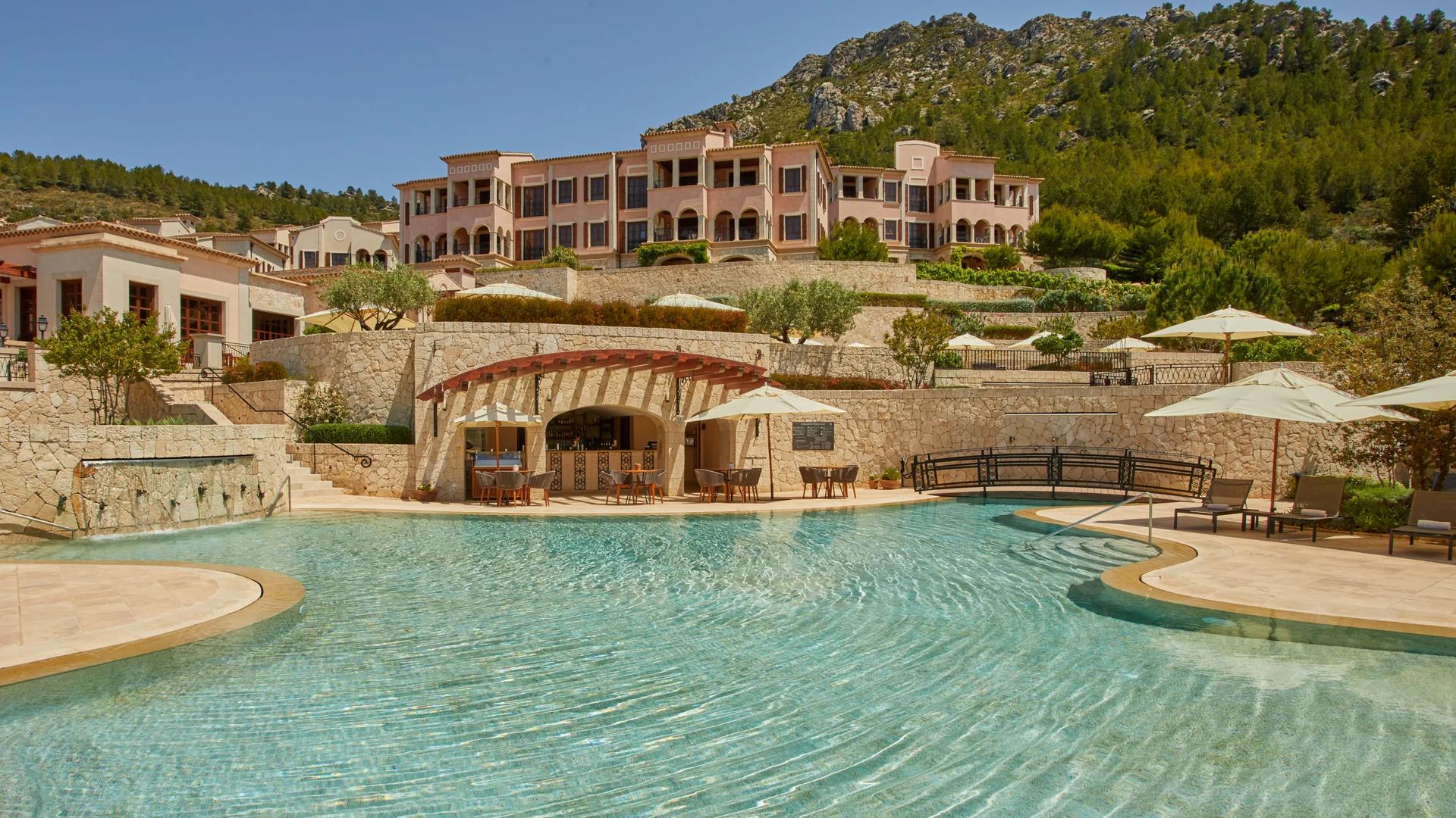 Large outdoor swimming pool at Vermell Grand Hotel in Mallorca, surrounded by stone terraces, lounge chairs, and light pink hillside buildings framed by forested mountains.