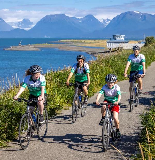 A family of four wearing matching green-and-white cycling jerseys and helmets riding bicycles along a paved coastal trail, with a body of water and snow-capped mountains in the background under a sunny sky.