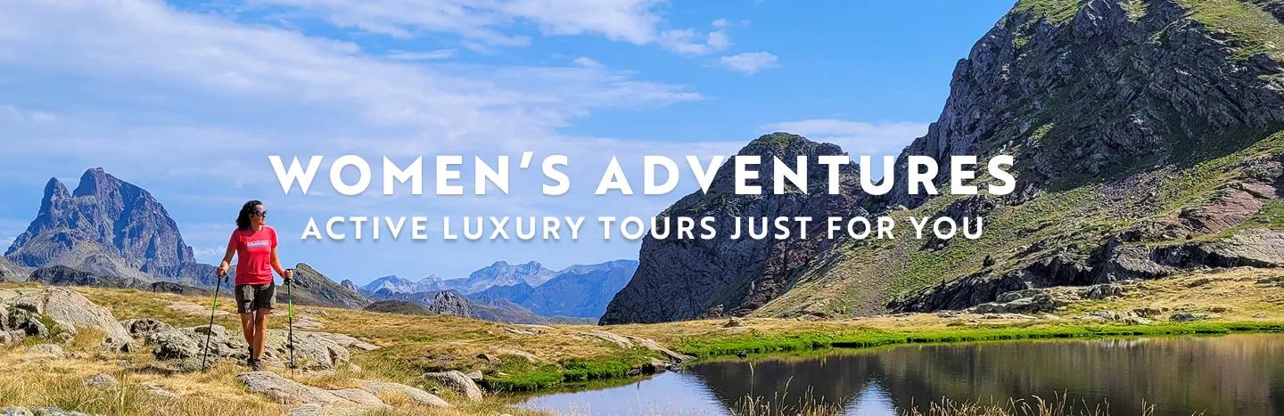 Panoramic landscape photo featuring a woman hiking with trekking poles along a grassy trail next to a calm lake, surrounded by rugged, rocky mountains under a partly cloudy blue sky. White overlay text in the center reads, "WOMEN'S ADVENTURES ACTIVE LUXURY TOURS JUST FOR YOU".