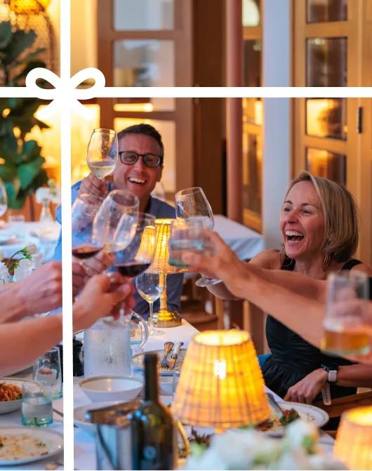 A group of people toasting and laughing together around a candlelit dining table. Image has a white bow overlay. 