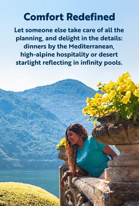 A woman with long dark hair, wearing a teal t-shirt, leans on a stone balcony railing with yellow flowers in a stone planter, looking out at a scenic lake surrounded by lush green mountains.   The text at the top reads:"Comfort RedefinedLet someone else take care of all the planning, and delight in the details: dinners by the Mediterranean, high-alpine hospitality or desert starlight reflecting in infinity pools."