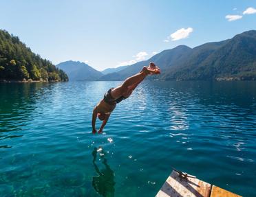 A person in mid-air diving headfirst into clear blue lake water from a wooden dock, with forested mountains surrounding the lake under a bright, sunny sky.