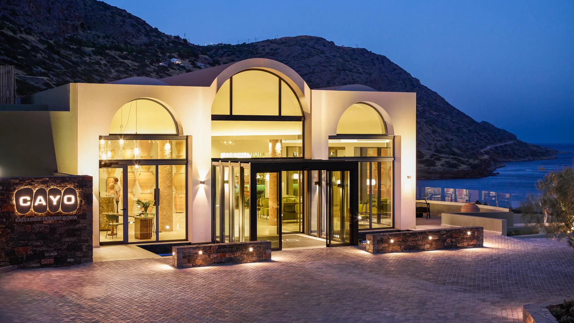 The Cayo Exclusive Resort & Spa in Plaka, in Crete, Greece features modern architecture with expansive glass windows and arched entrances overlooking the sea.