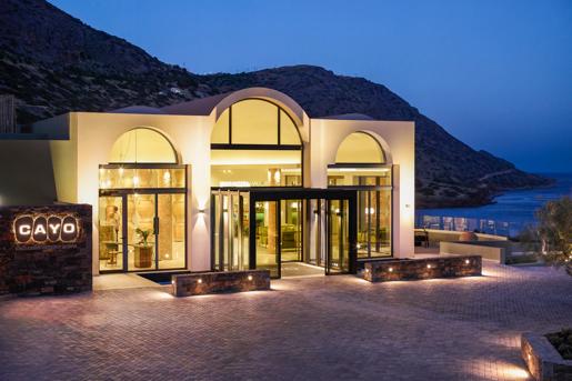 The Cayo Exclusive Resort & Spa in Plaka, in Crete, Greece features modern architecture with expansive glass windows and arched entrances overlooking the sea.