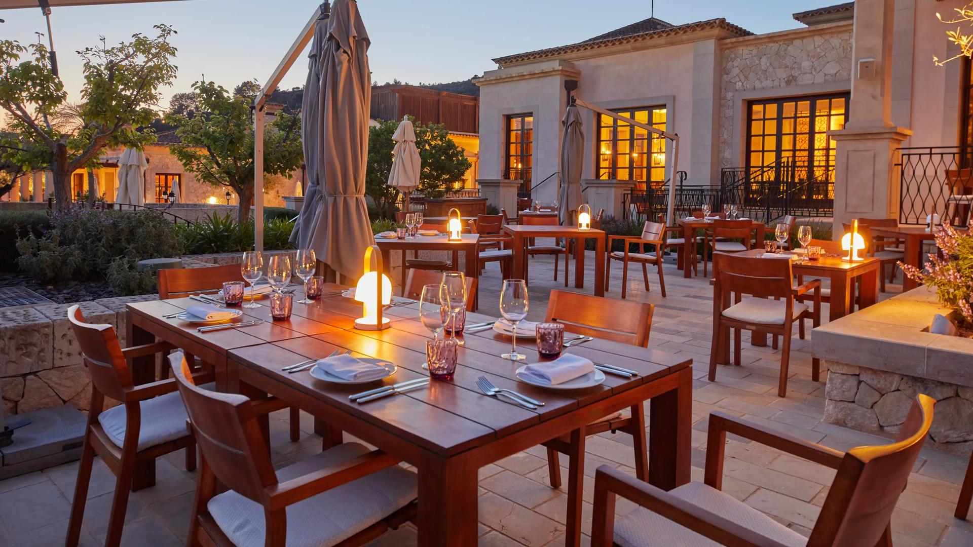 Twilight view of Cap Vermell Grand Hotel dining terrace set with wooden tables, warm table lamps, and fine glassware beside Mediterranean villa buildings.
