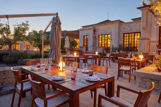 Twilight view of Cap Vermell Grand Hotel dining terrace set with wooden tables, warm table lamps, and fine glassware beside Mediterranean villa buildings.
