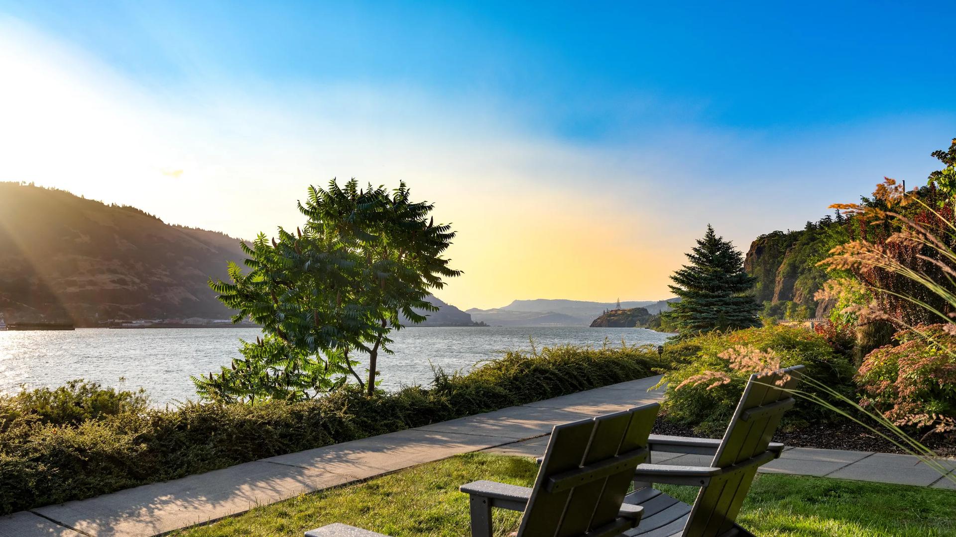 Two dark wooden Adirondack chairs sit on a green lawn facing a scenic river surrounded by mountains at sunset.