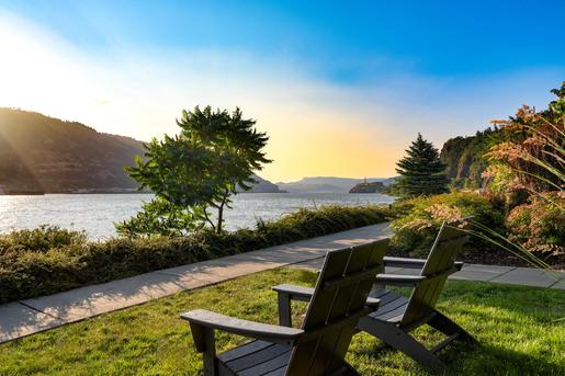 Two dark wooden Adirondack chairs sit on a green lawn facing a scenic river surrounded by mountains at sunset.