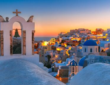 Sunset view of Oia, Santorini, featuring a traditional white bell tower and blue-domed churches cascading down a cliffside by the sea.