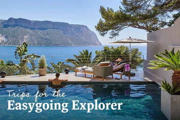 A couple relaxes in an upscale outdoor pool area overlooking a wide blue bay with a massive, sheer mountain cliff rising from the water in the background. Text overlay reads: Trips for the Easygoing Explorer. 