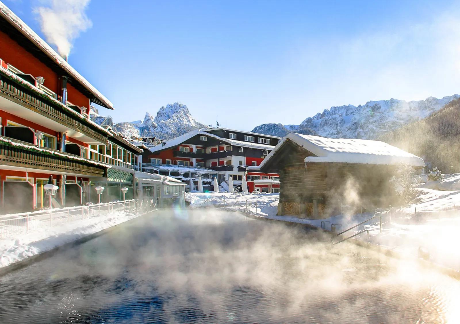 The heated outdoor pool with rising steam in winter, set against the traditional Alpine chalet architecture at the Alpenroyal Hotel in Selva di Val Gardena, Italy.