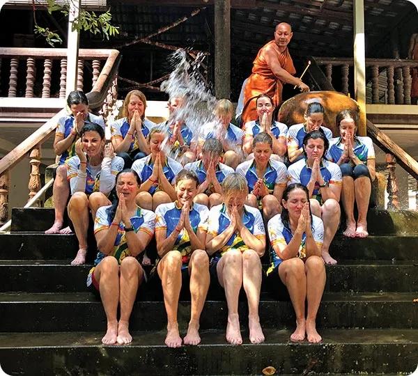 Here is alt text for this image:

**Alt text:** A group of women in matching athletic shirts sitting on outdoor wooden stairs with hands pressed together in a prayer position, as a Buddhist monk in orange robes pours water over them during a blessing ceremony.