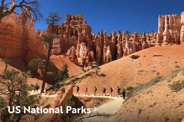 A line of hikers walking along a dirt trail surrounded by tall red rock hoodoos and cliffs under a clear blue sky, with text reading "US National Parks" overlaid in the bottom left corner.