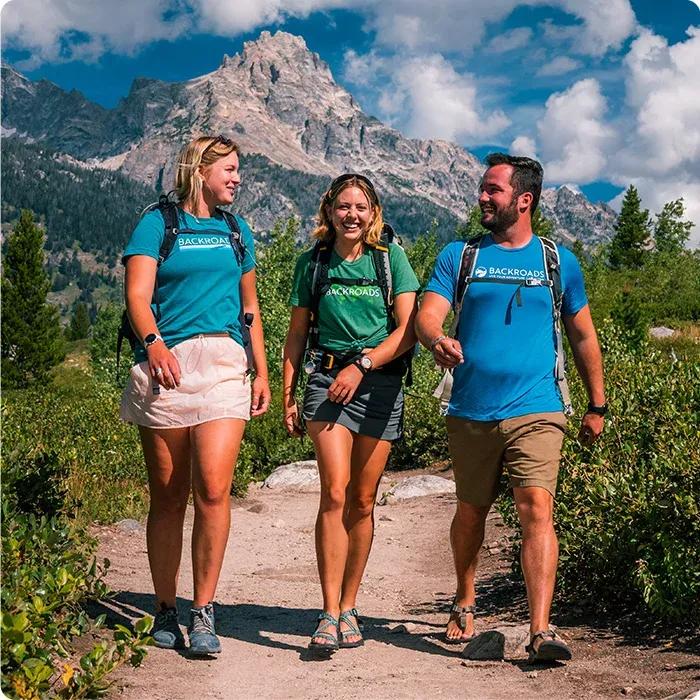 Three smiling hikers wearing backpacks and Backroads shirts walking together along a dirt trail with dramatic, rugged mountain peaks in the background under a blue sky.