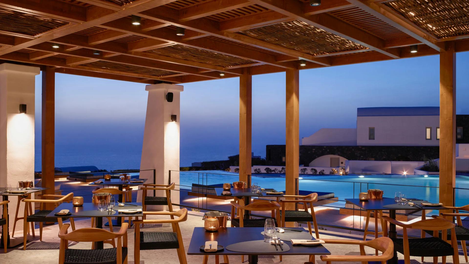 The Alios Ilios restaurant in Santorini, Greece, features a covered outdoor dining terrace overlooking an infinity pool and the Aegean Sea.