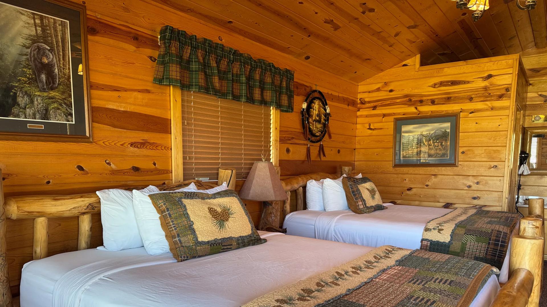 The double-queen rustic log interior of Badlands Frontier Cabins in Wall, South Dakota with Cabin-themed patchwork blankets, pine cone accent pillows, wood-paneled walls, and nature artwork.