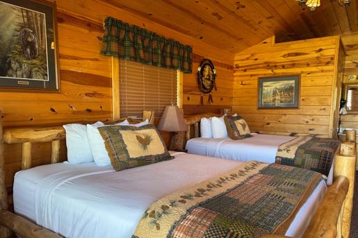 The double-queen rustic log interior of Badlands Frontier Cabins in Wall, South Dakota with Cabin-themed patchwork blankets, pine cone accent pillows, wood-paneled walls, and nature artwork.