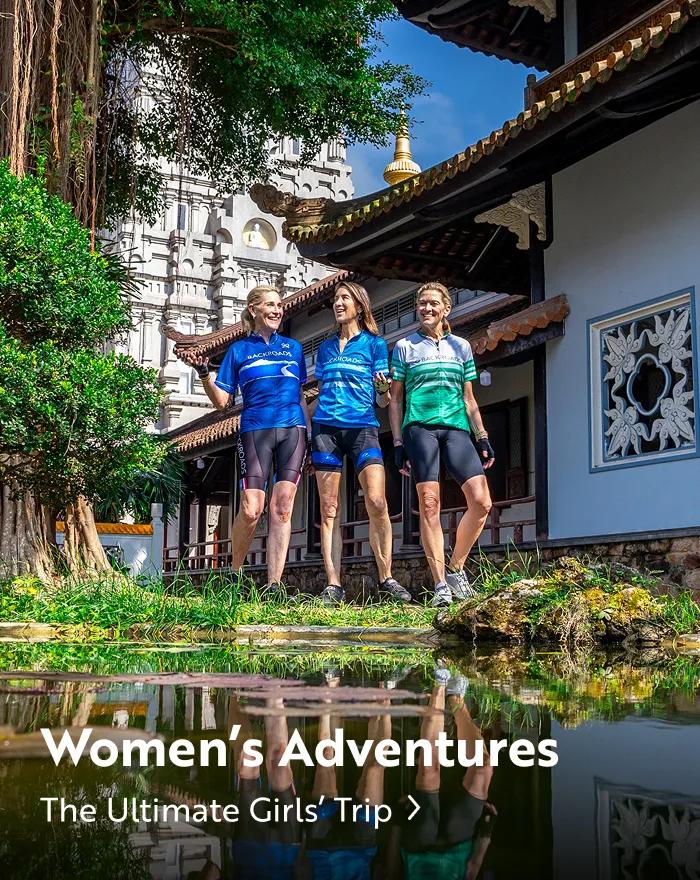 Three women in cycling gear smile together in front of a traditional temple with a calm pond in the foreground. Text overlays the bottom reading: "Women's Adventures. The Ultimate Girls' Trip".