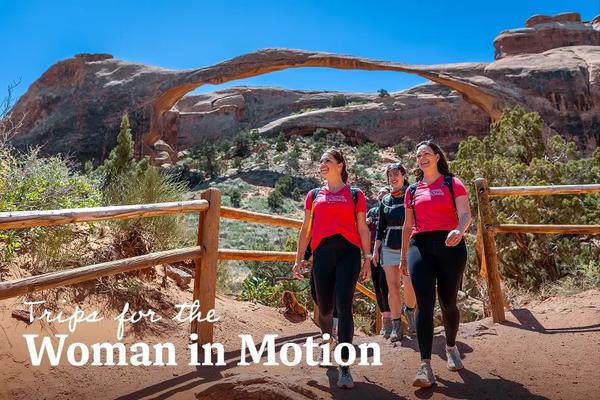 A group of women hiking past blooming red cactus flowers near the towering sandstone spires near Arches and Canyonlands National Parks, Utah. Text overlay reads: Trips for the Woman in Motion.
