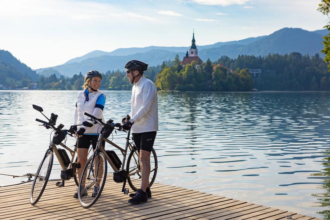 Two Backroads cyclists pause at a lake with a church spire in the background in Slovenia.