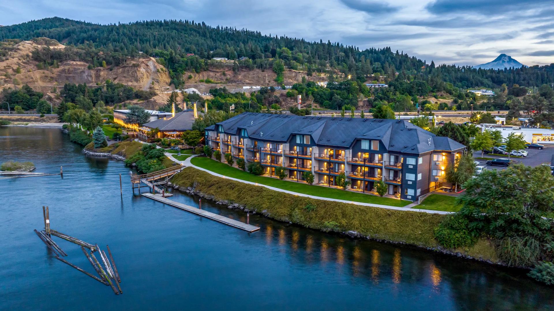 Aerial view of the Hood river Inn at the Hood river in Oregon, situated along a calm river with lush green hills and a snow-capped mountain peak in the background at twilight.   