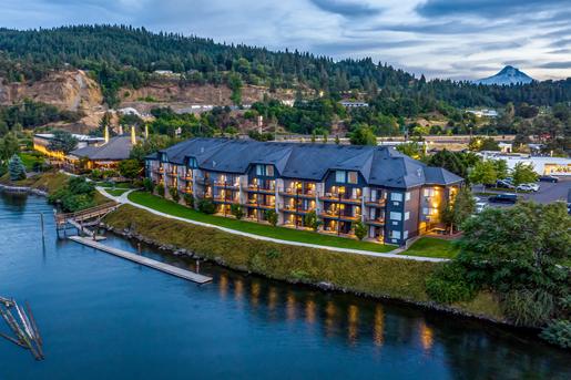 Aerial view of the Hood river Inn at the Hood river in Oregon, situated along a calm river with lush green hills and a snow-capped mountain peak in the background at twilight.