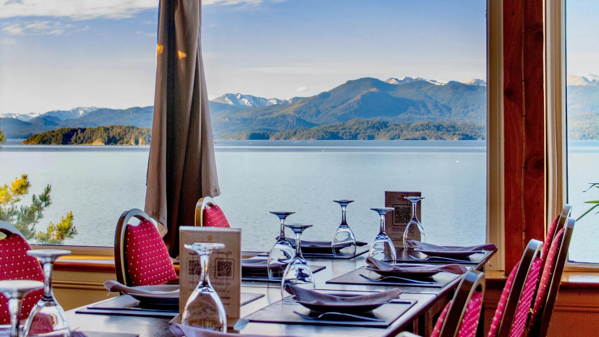 An indoor restaurant dining table set with wine glasses, placemats, and red chairs next to large picture windows at Sol del Nahuel Hotel & Spa in San Carlos de Bariloche, Argentina, providing expansive views of Lake Nahuel Huapi and distant mountains under a partly cloudy sky.