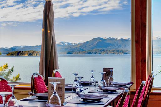 An indoor restaurant dining table set with wine glasses, placemats, and red chairs next to large picture windows at Sol del Nahuel Hotel & Spa in San Carlos de Bariloche, Argentina, providing expansive views of Lake Nahuel Huapi and distant mountains under a partly cloudy sky.